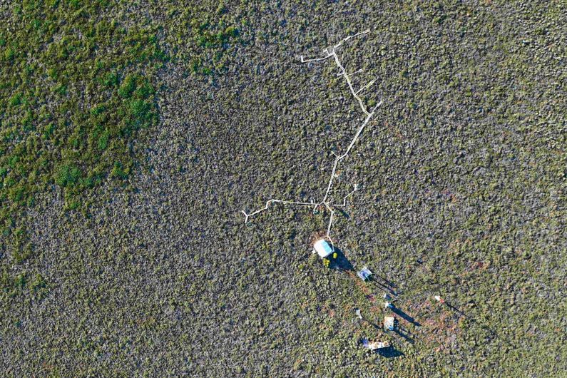 Vue aérienne prise par drone en juin 2019 de la station de recherche Trail Valley Creek, dans les Territoires du Nord-Ouest, où des mesures automatisées et manuelles de flux de gaz sont effectuées. Le trottoir de bois permet d'accéder à chaque chambre de mesure sans nuire à la végétation ni altérer les flux de gaz à l'intérieur des chambres. La tente blanche abrite l'analyseur de gaz et le dispositif de contrôle du système de chambres automatisées. Une station micrométéorologique et une tour de covariance des turbulences sont visibles au bas de la photo.